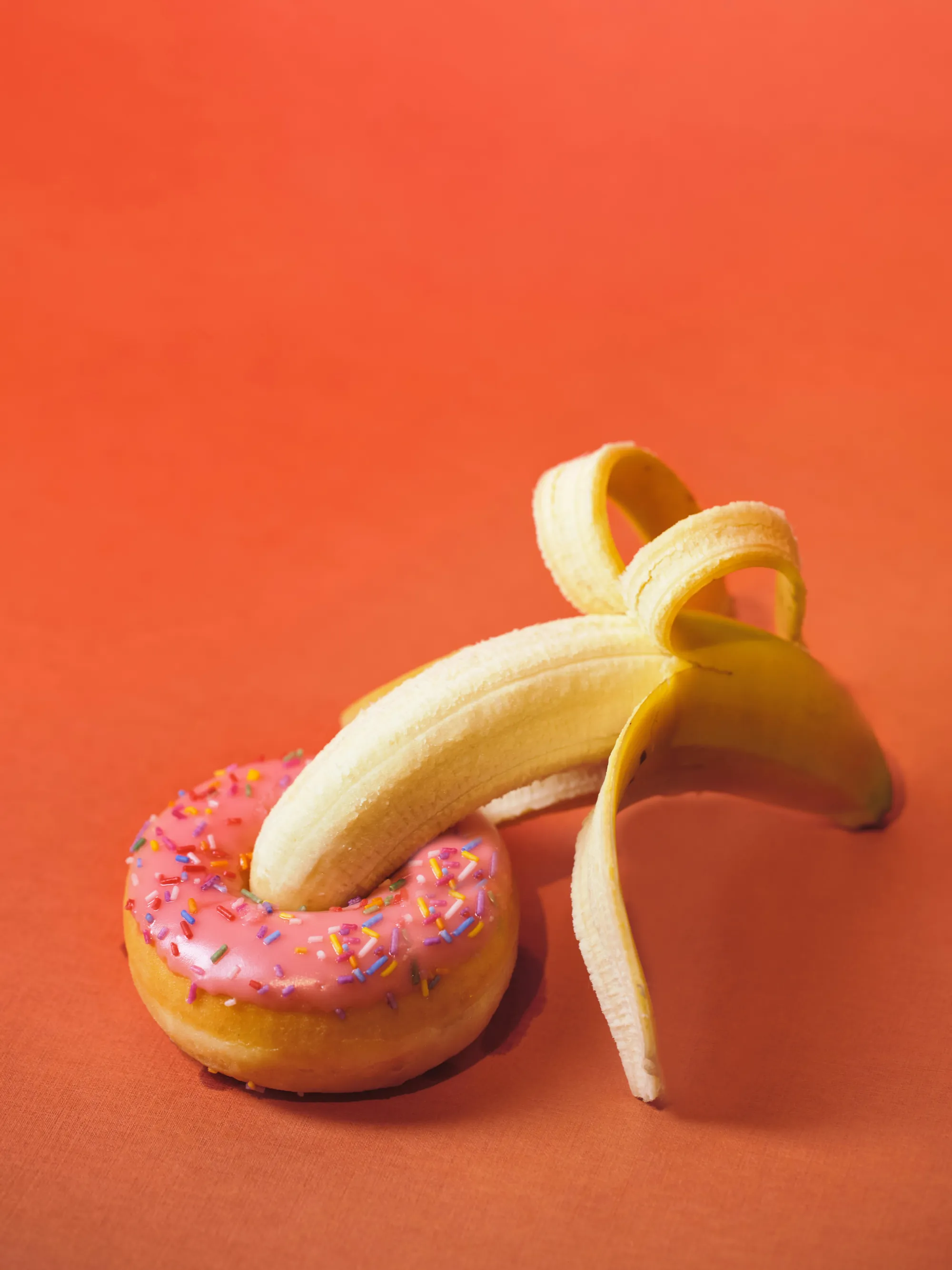 Eine geschälte Banane, gesteckt durch einen rosa Donut mit Streuseln
